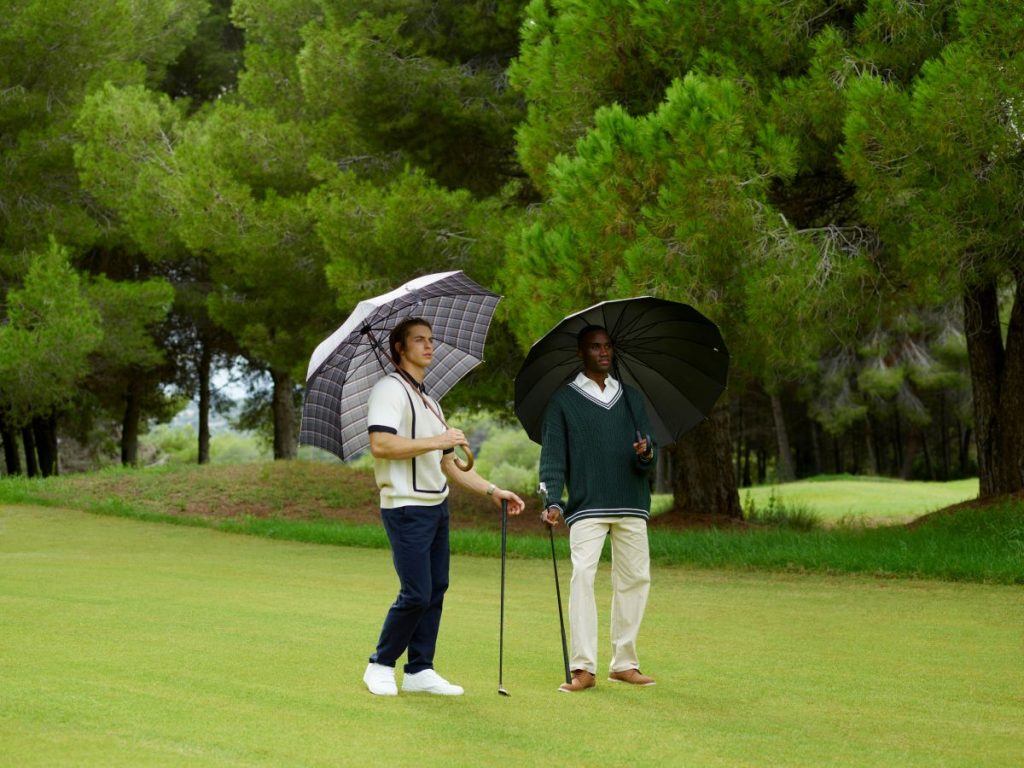 Two golfers walk across a green holding clubs and a logo golf umbrella on a tree-lined course.