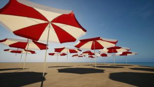 Red and white striped promotional umbrellas arranged on a sunny beach, with a clear blue sky and calm ocean in the background.
