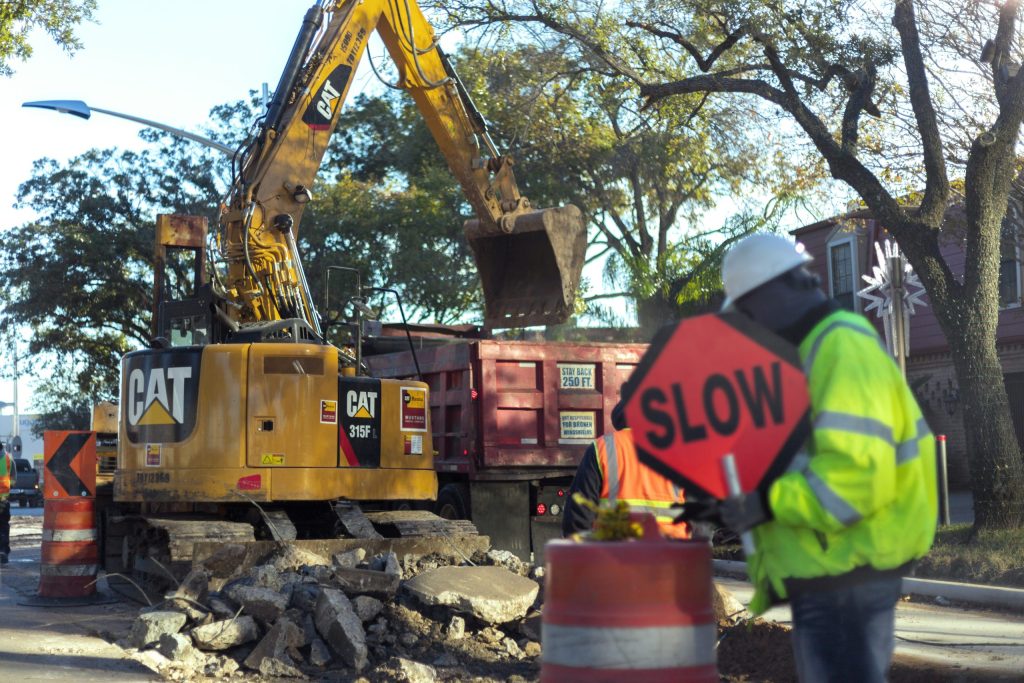 Construction site with a yellow CAT excavator loading debris into a red dump truck, workers directing traffic with a 'SLOW' sign, and rubble on the ground, illustrating groundwork preparation for a California Bearing Ratio (CBR) test