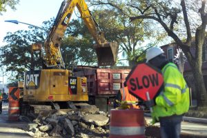 Construction site with a yellow CAT excavator loading debris into a red dump truck, workers directing traffic with a 'SLOW' sign, and rubble on the ground, illustrating groundwork preparation for a California Bearing Ratio (CBR) test
